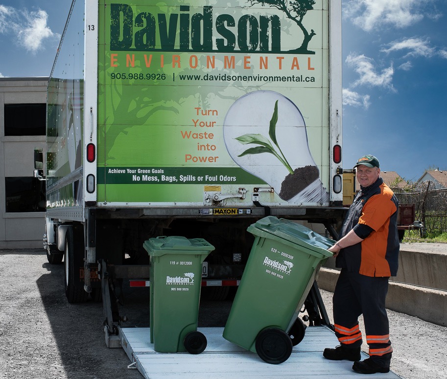 A worker stands next to two green waste bins, smiling as he prepares to load them onto a Davidson Environmental truck, promoting waste-to-energy services.