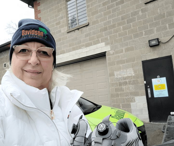 Woman in a white jacket and blue beanie smiles while holding cycling shoes outside a garage, with a bright green car in the background.