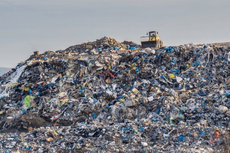 A large landfill mound covered in mixed waste, including plastic, appliances, and debris, with a small structure visible at the summit.