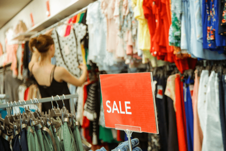 A shopper selects clothes from a rack in a retail store, with a prominent "SALE" sign in the foreground, highlighting a discount event.