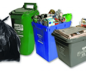 Three recycling bins and a garbage bag arranged side by side. The green bin is for organics, the blue for recyclables, and the gray for paper waste.