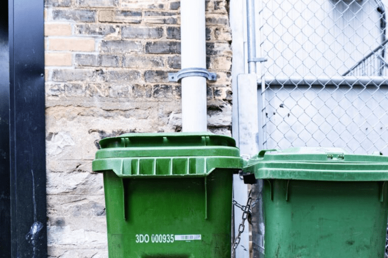 Two green trash bins are positioned against a brick wall, chained together, showcasing urban waste management in a compact space.