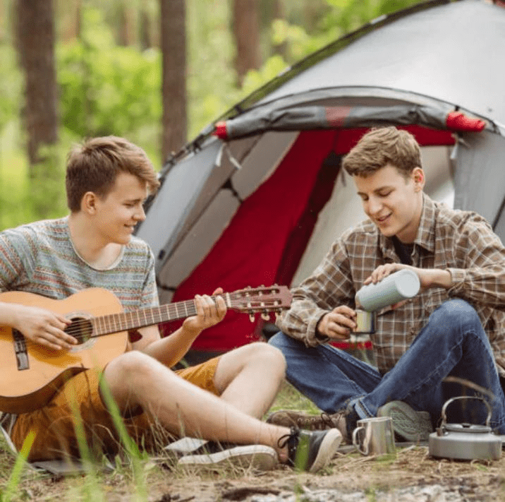 Teen boys enjoy a camping trip, with one playing guitar while the other pours from a thermos, surrounded by nature and a tent.