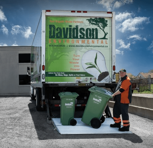 A waste management worker, wearing an orange and black uniform, rolls two green bins from a Davidson Environmental truck in a recycle facility.