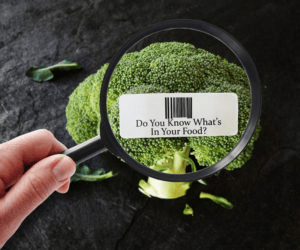 A hand holding a magnifying glass over a piece of broccoli displaying a label that asks, "Do You Know What's In Your Food?" This highlights food transparency.