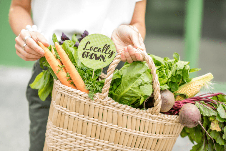 A person holds a woven basket filled with fresh, vibrant produce, including carrots and leafy greens, with a "Locally GROWN" sign prominently displayed.